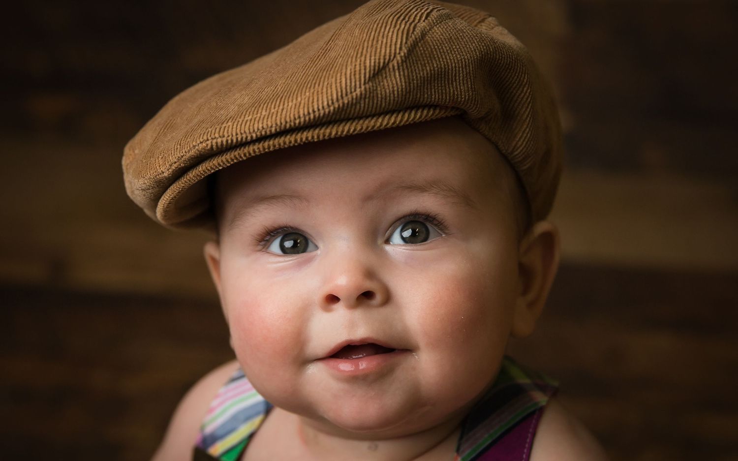 Baby wearing brown newsboy cap smiles at camera in studio portrait.