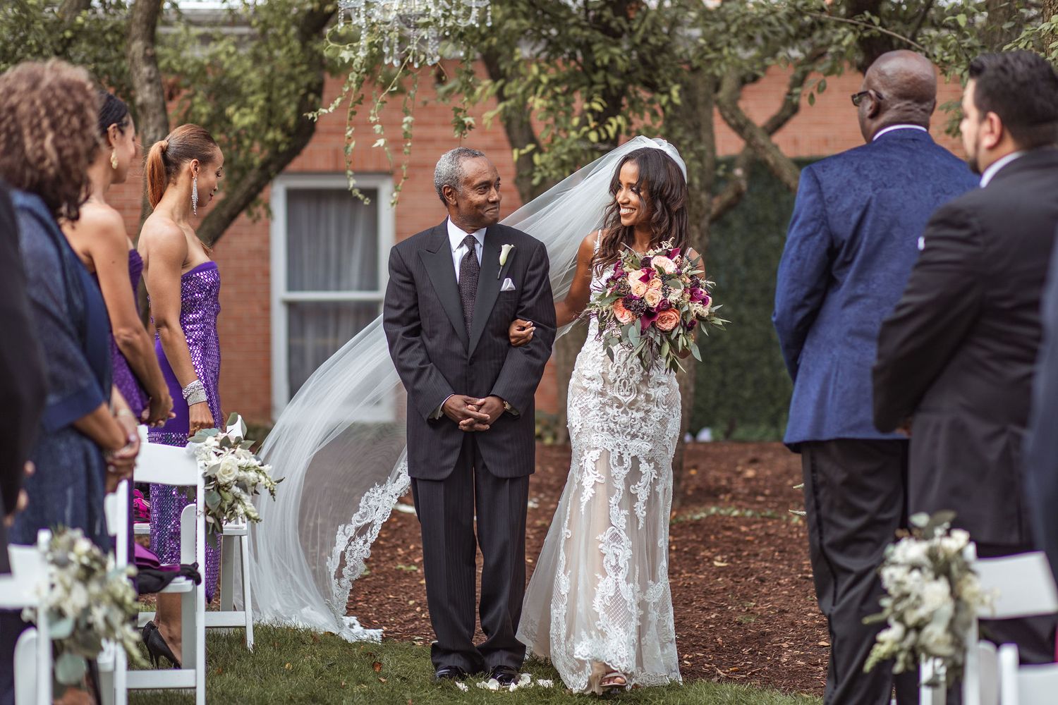 An outdoor wedding ceremony under trees with purple-clad attendants and floral decorations.