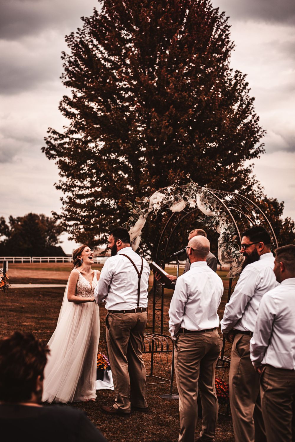 An outdoor wedding ceremony takes place under a large tree with a rustic arch and bridal party in casual attire.