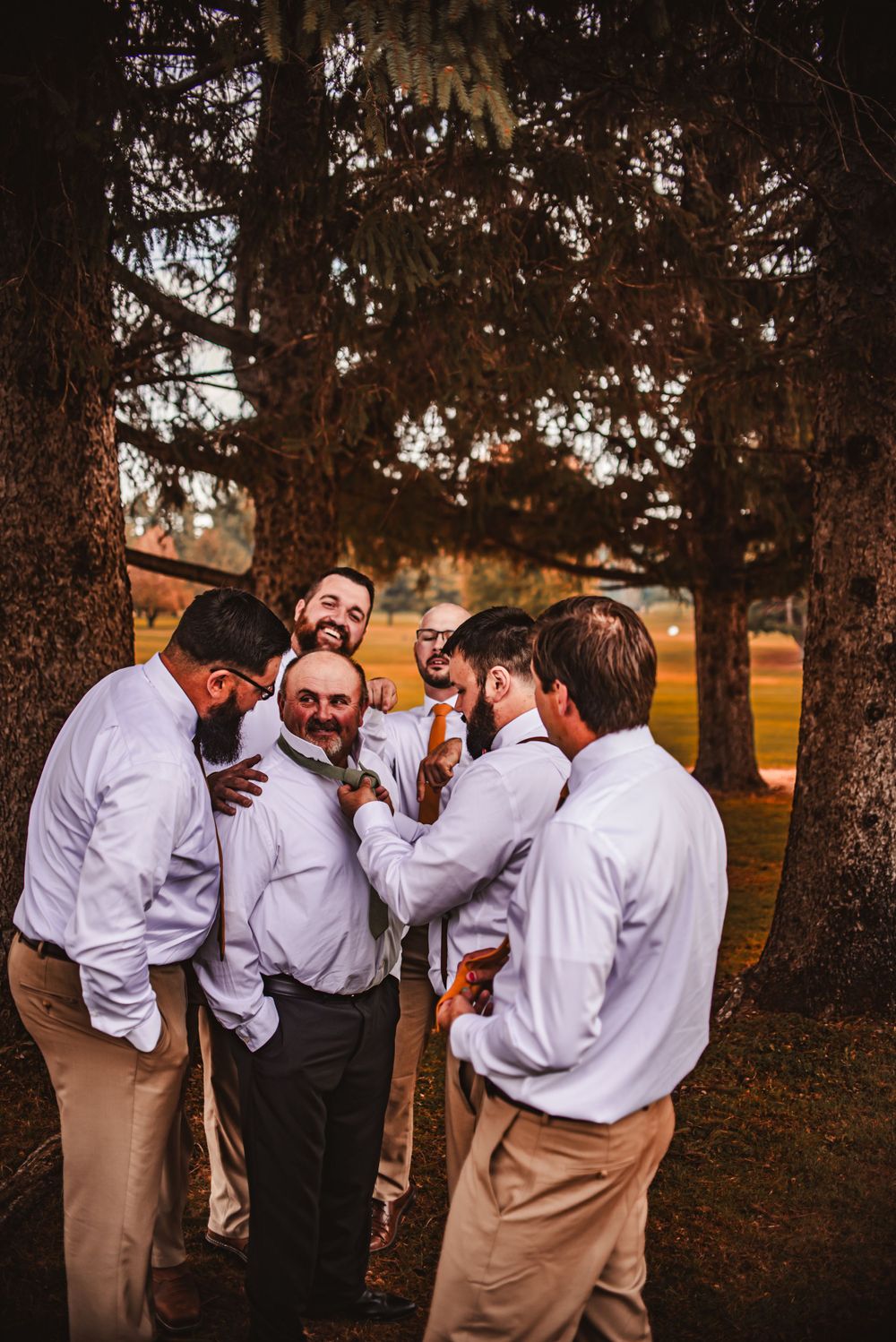A group of groomsmen in white shirts and khakis share a lighthearted moment under trees at sunset.