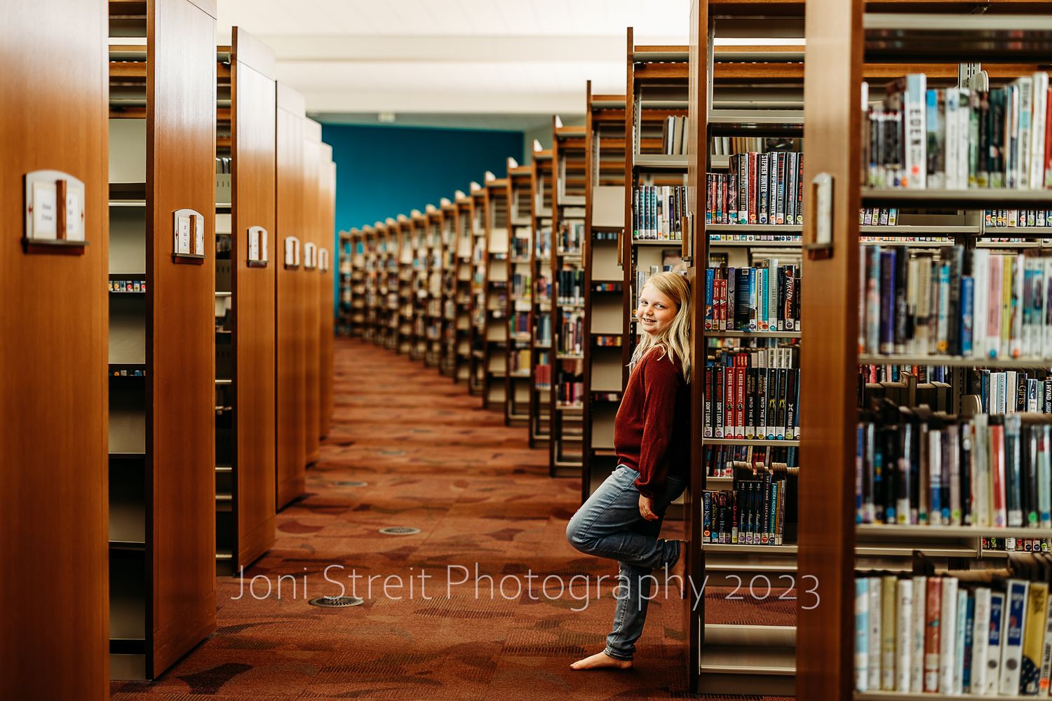 Long library aisle with wooden shelving and books stretches into the distance beneath turquoise walls.