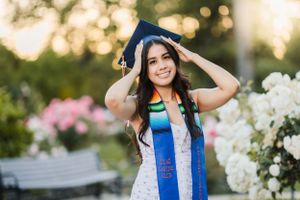 Graduate in white dress and blue stole poses with flowers in garden setting.