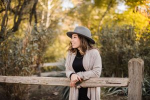 A figure in a gray hat and sweater leans thoughtfully against a wooden fence in an autumn setting.
