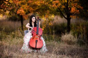 A musician plays cello outdoors in a white dress surrounded by autumn foliage.