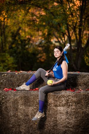 An athlete in blue and gray sportswear sits on a stone wall with baseball equipment.