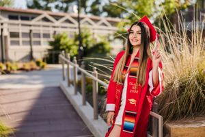 A graduate in red academic regalia stands on a modern campus walkway.