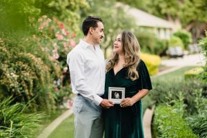 Couple shares intimate moment in garden setting while holding ultrasound photo.