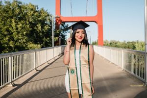 Graduate in white and green regalia poses on red bridge during sunny day.