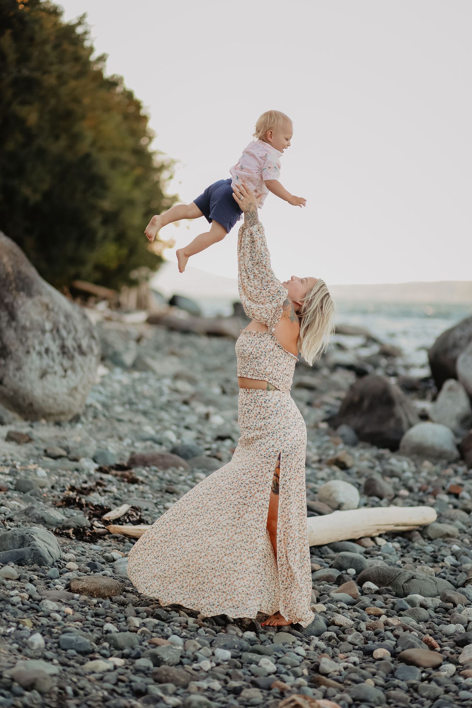 Mother and child playing on rocky beach in flowing floral dress.