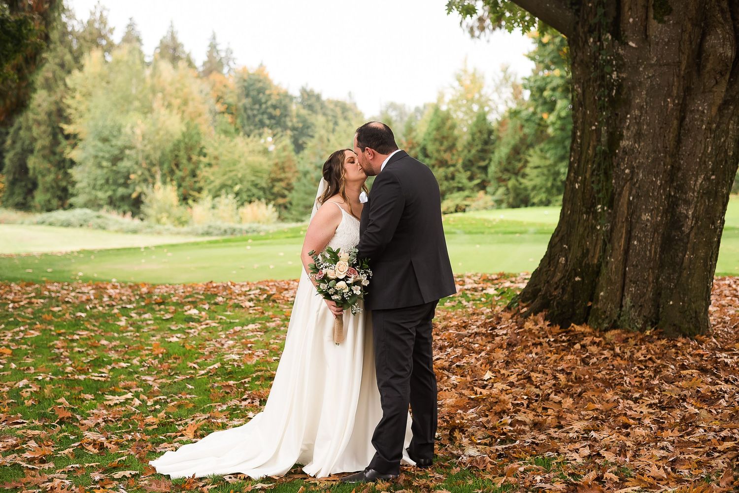 Standing amongst the colourful autumn leaves, a bride and groom kiss while she holds her wedding bouquet.