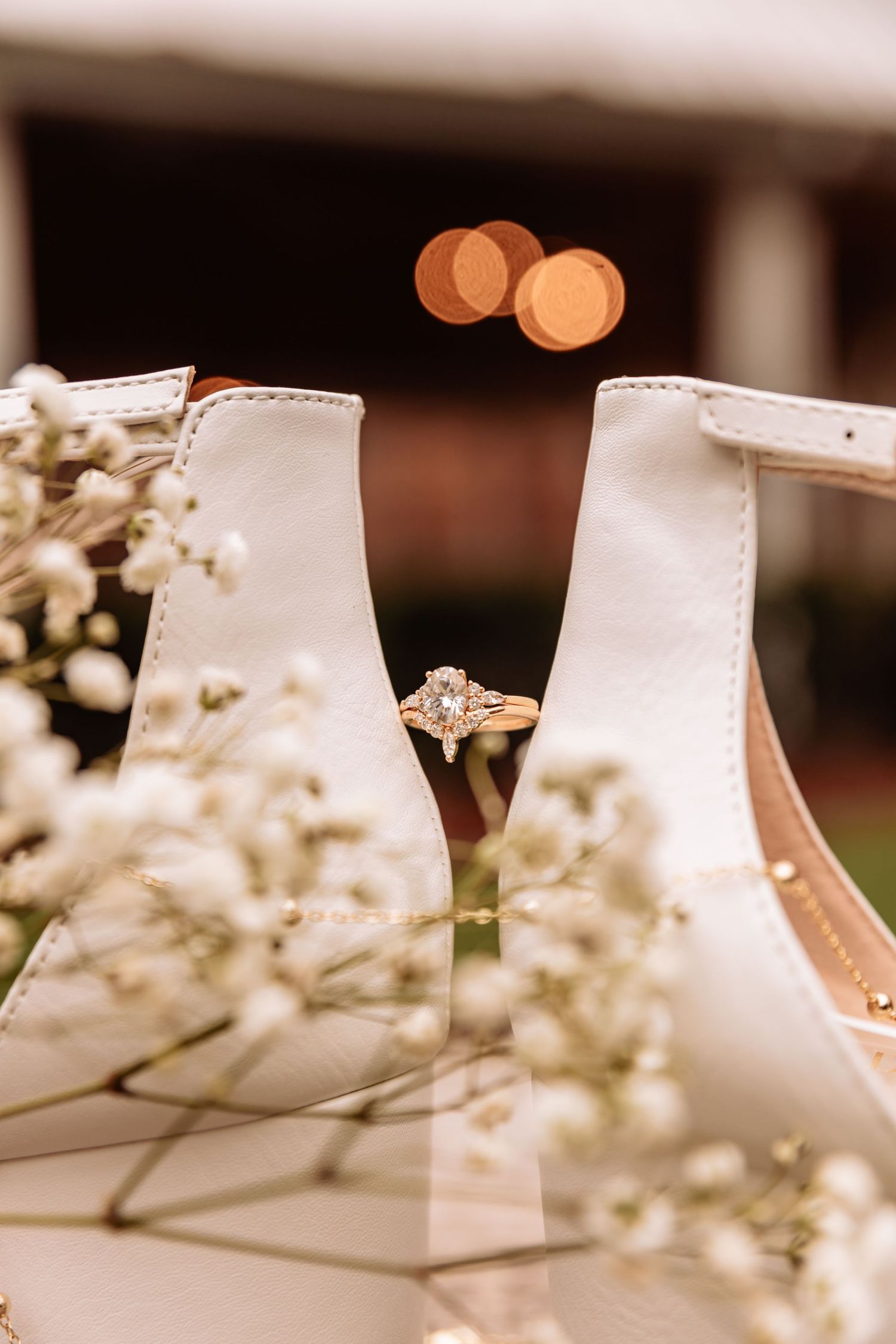Wedding ring sparkles against soft bokeh lights with delicate white flowers in the foreground.