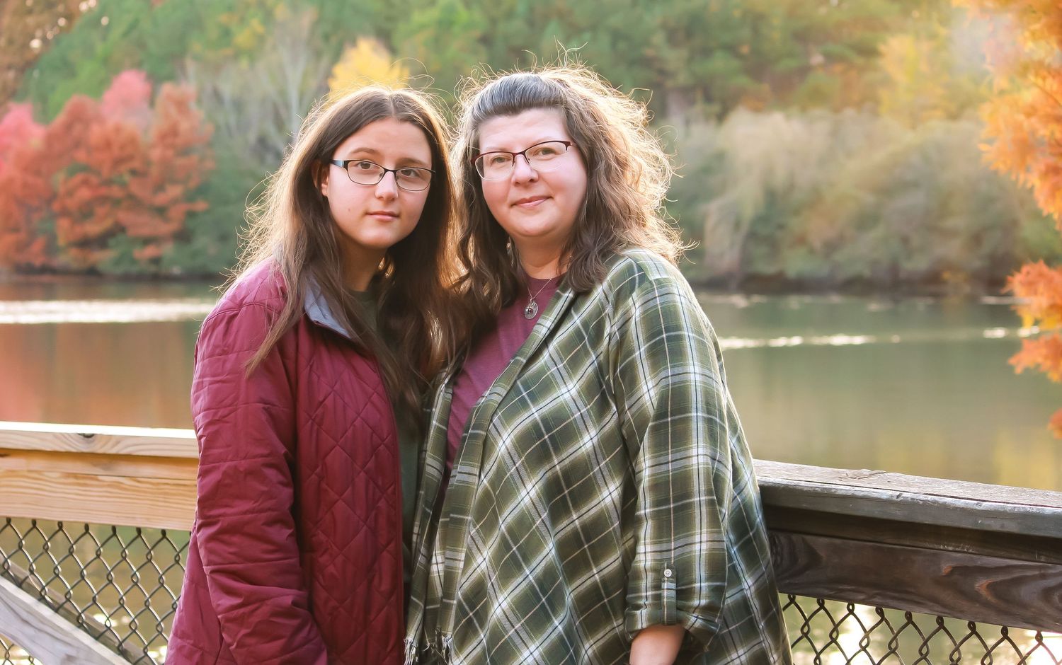 Two friends share a moment on bridge deck with colorful fall foliage and lake behind them.