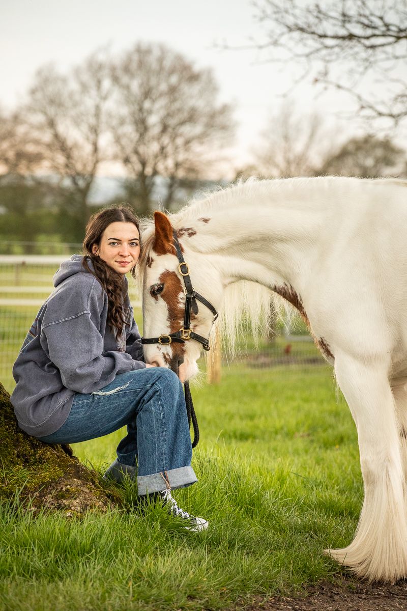 A person in casual wear crouches beside a white horse in a grassy field during sunset.