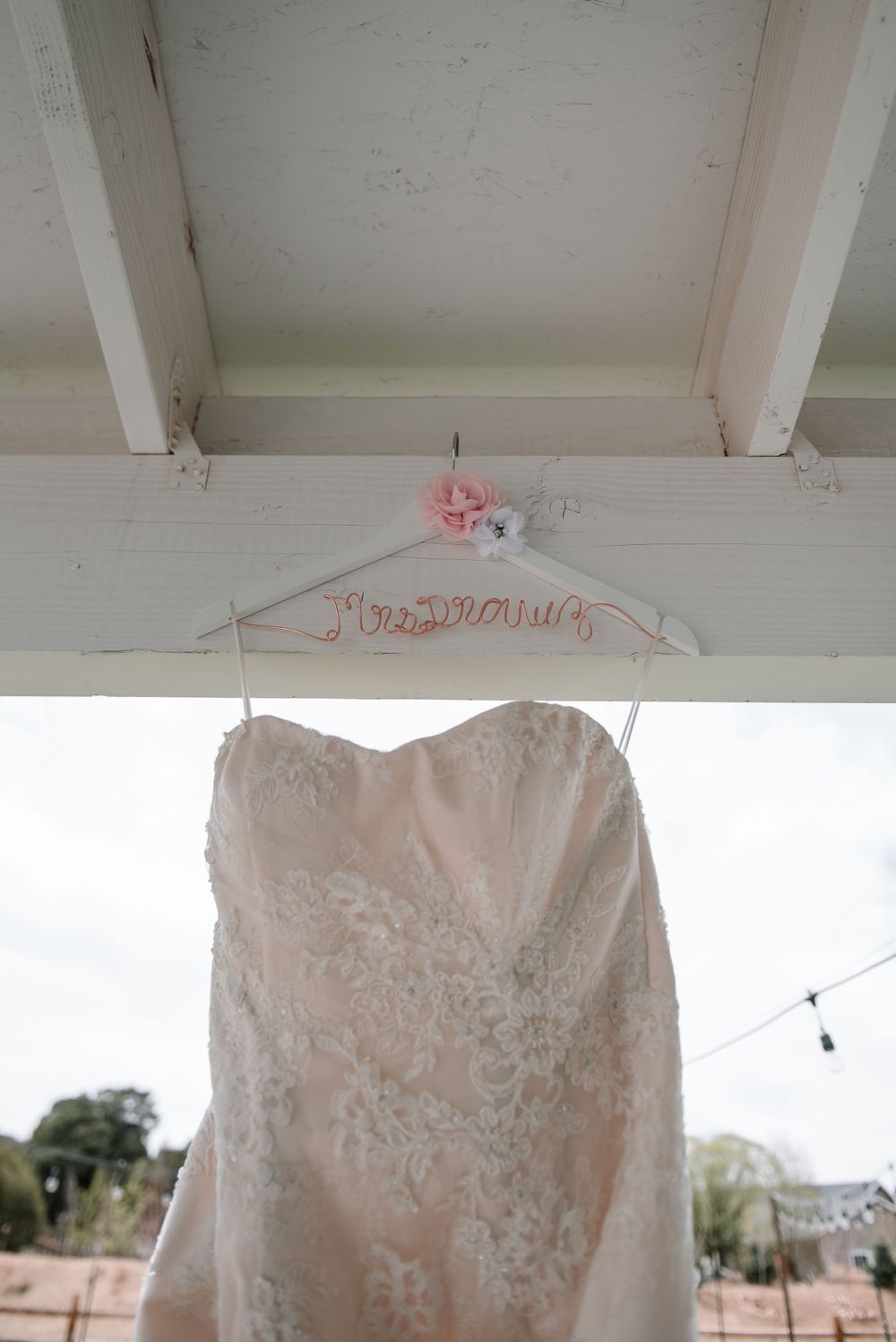 White lace wedding dress hanging from a decorative hanger against a light-colored ceiling.