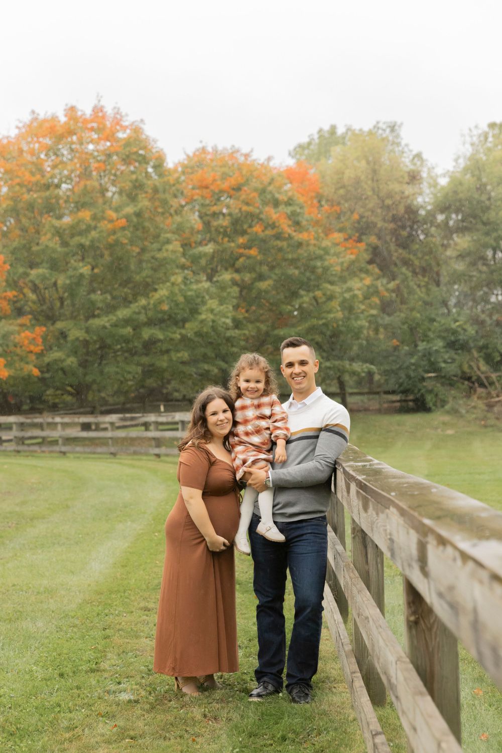 family of three with a pregnant mom smile for a photo with a wooden fence and orange trees