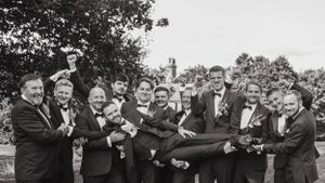 Black and white photo of groomsmen lifting someone in formal attire during outdoor celebration.