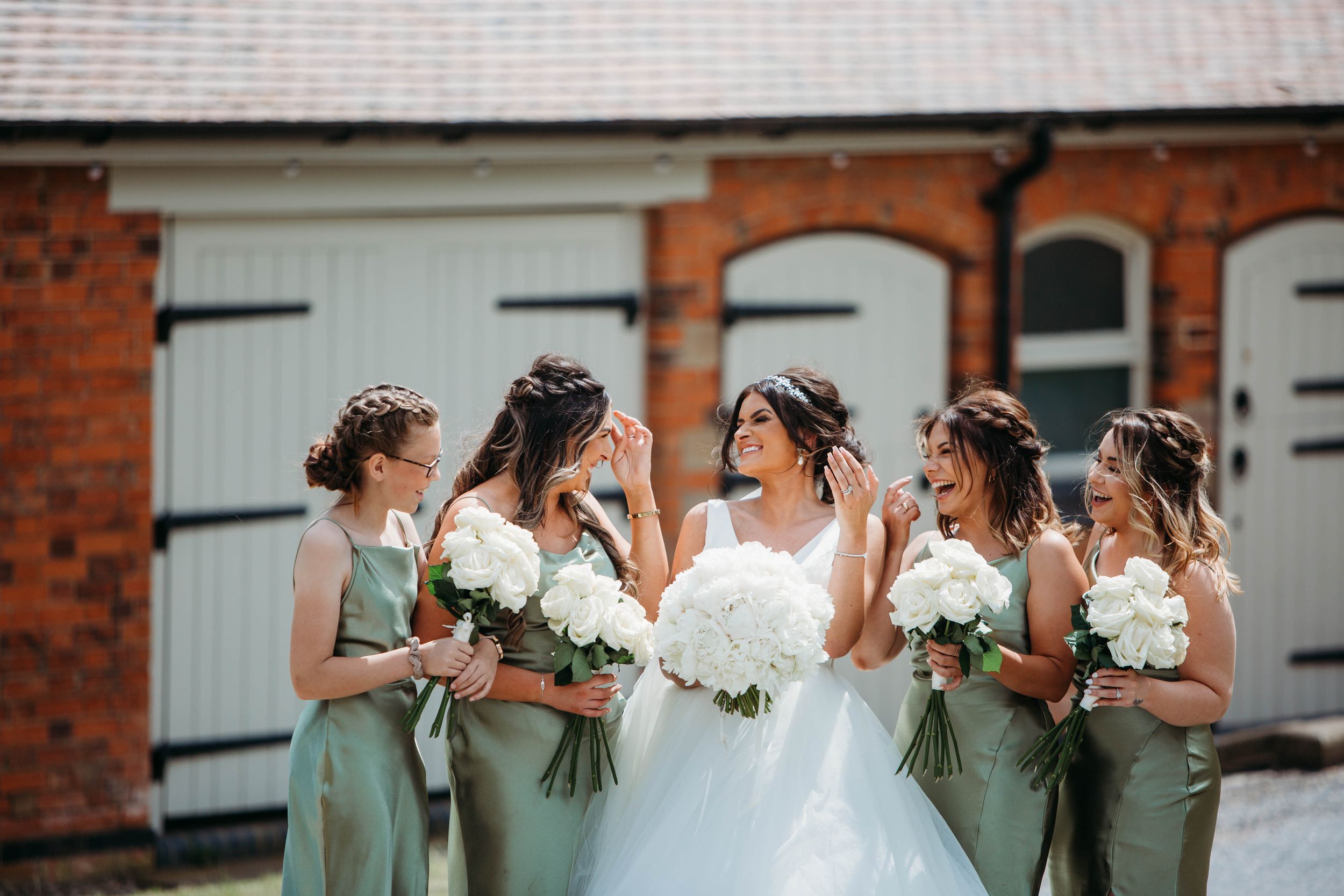 Bridesmaids in sage green dresses share a moment with bride in front of brick building.