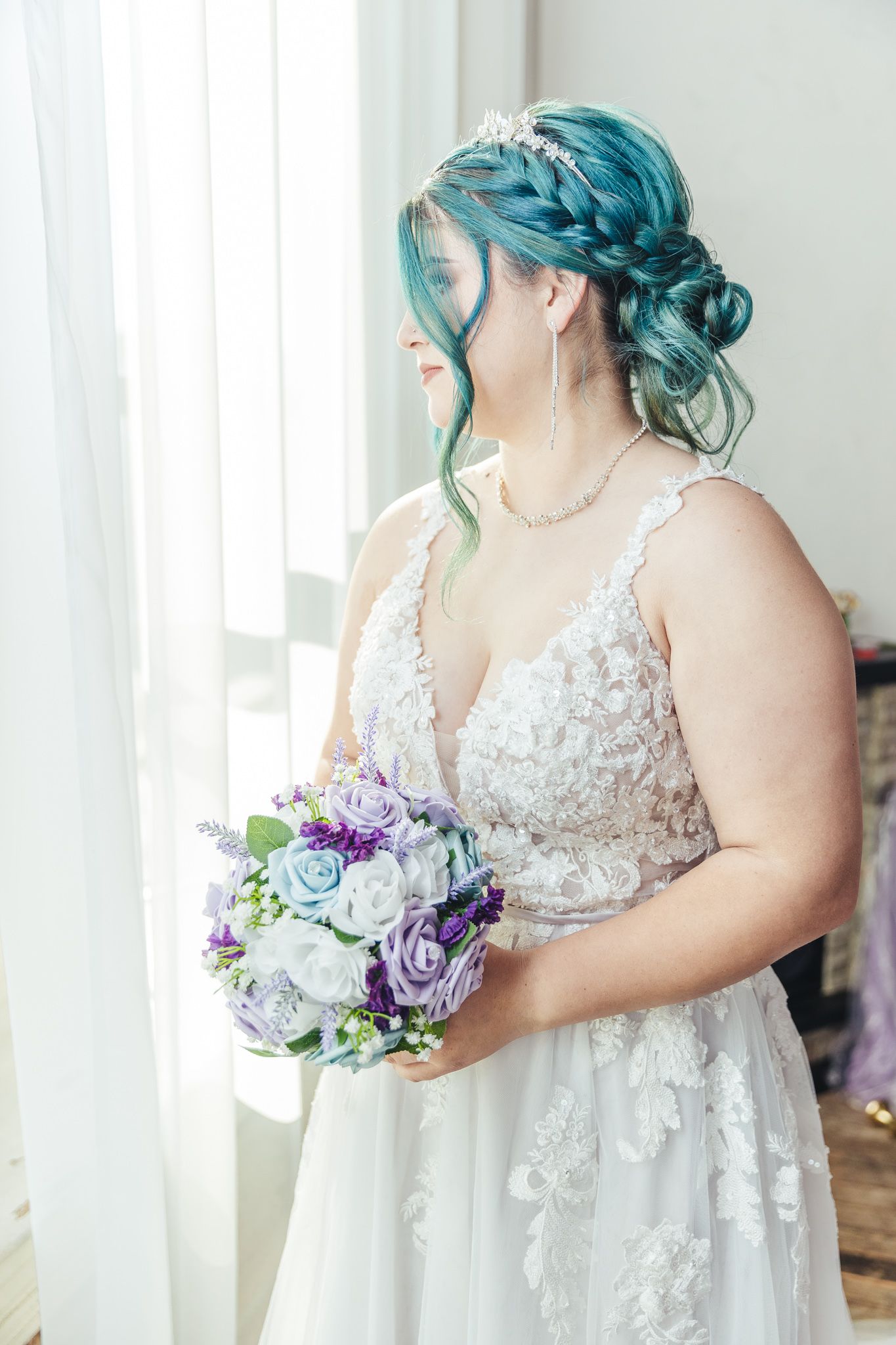 Bride in lace gown holds purple and white bouquet against white backdrop.