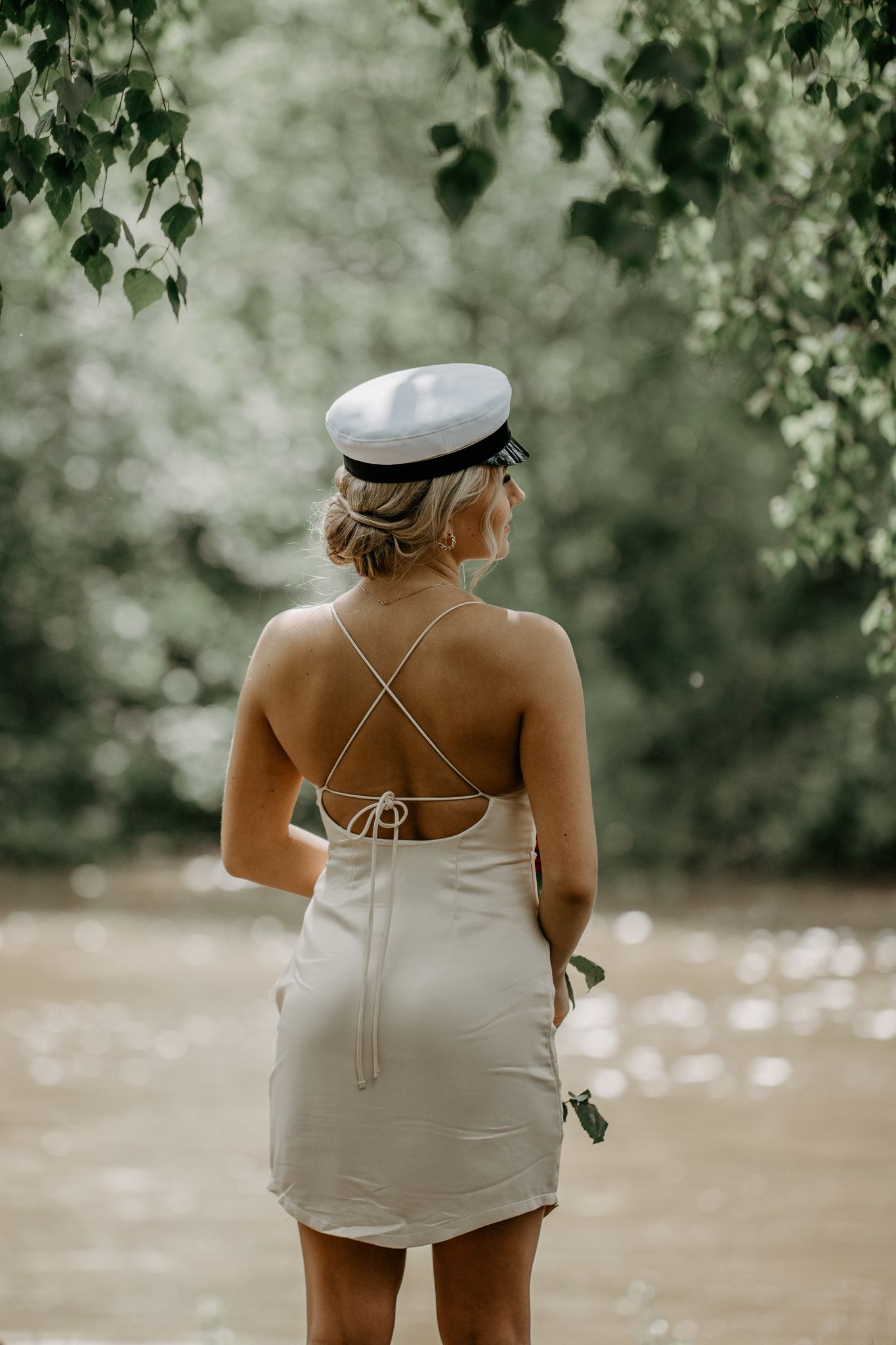Figure in white dress and sailor cap stands near water with greenery overhead.
