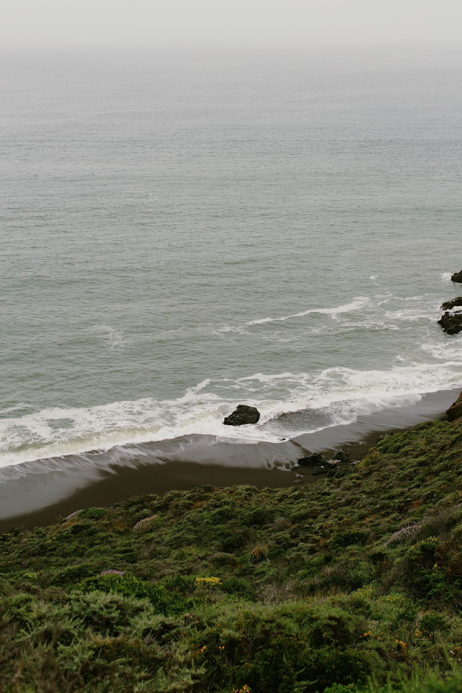 Dramatic coastal view of waves crashing against a black sand beach below green cliffs.