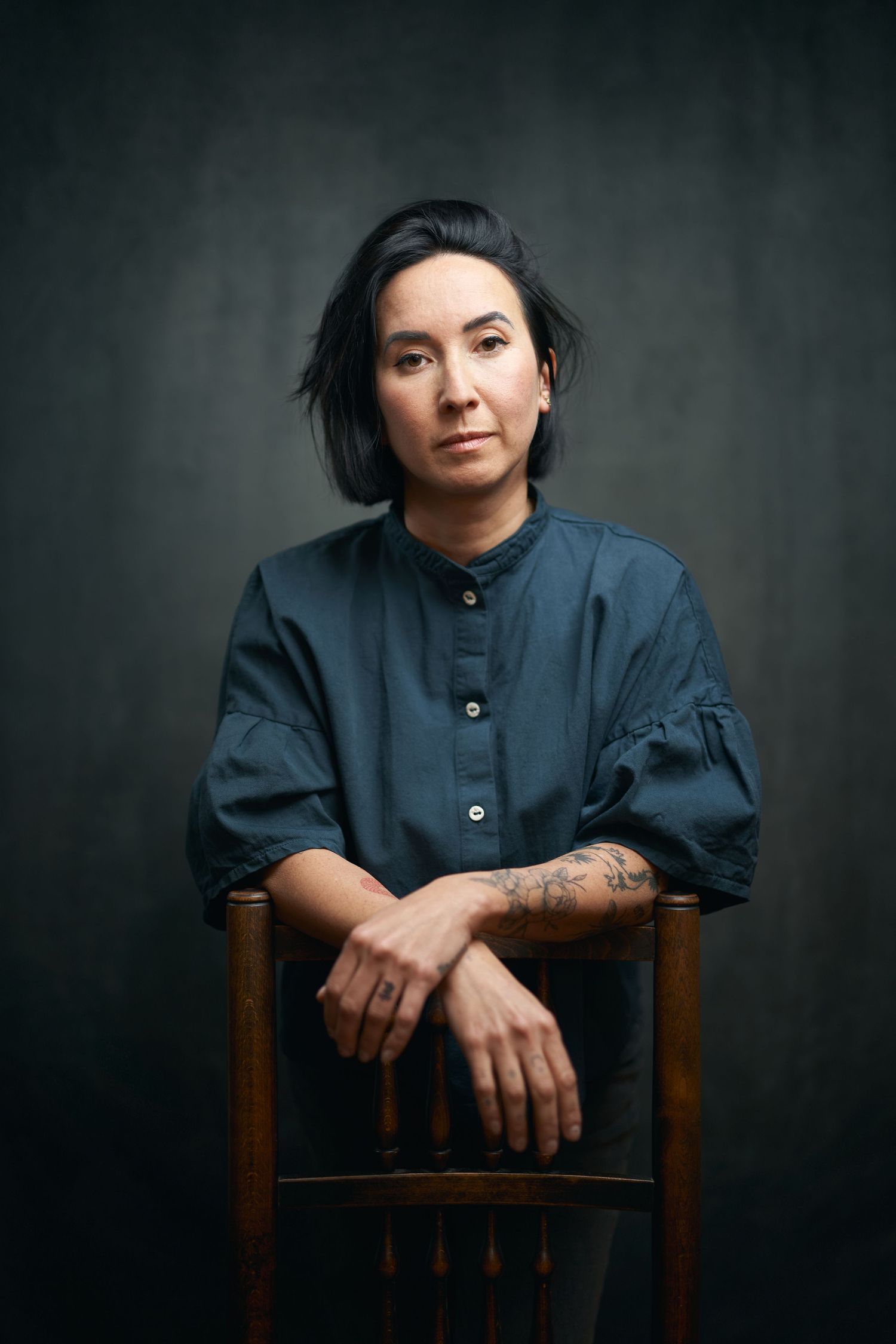 Studio portrait against dark background showing person in blue button-up shirt seated on wooden chair.