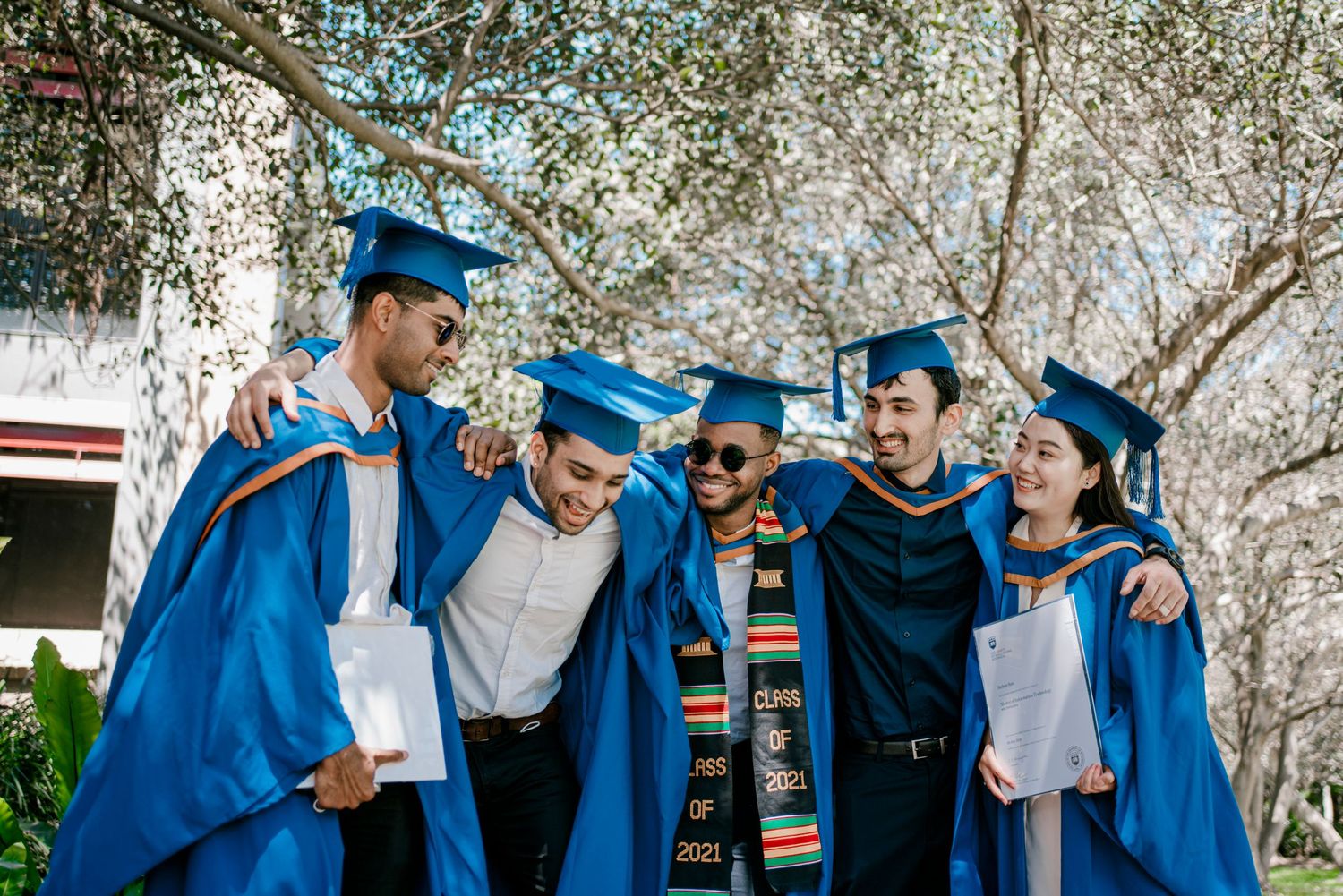 Group of graduates in blue robes smiling and posing together on a sunny day.