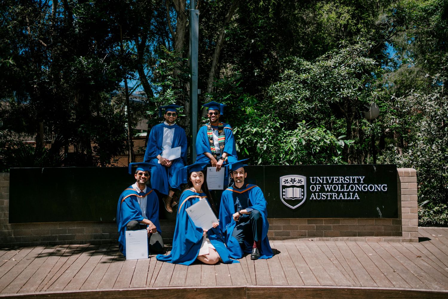 Group of graduates in blue gowns sitting casually on wooden deck during outdoor celebration.