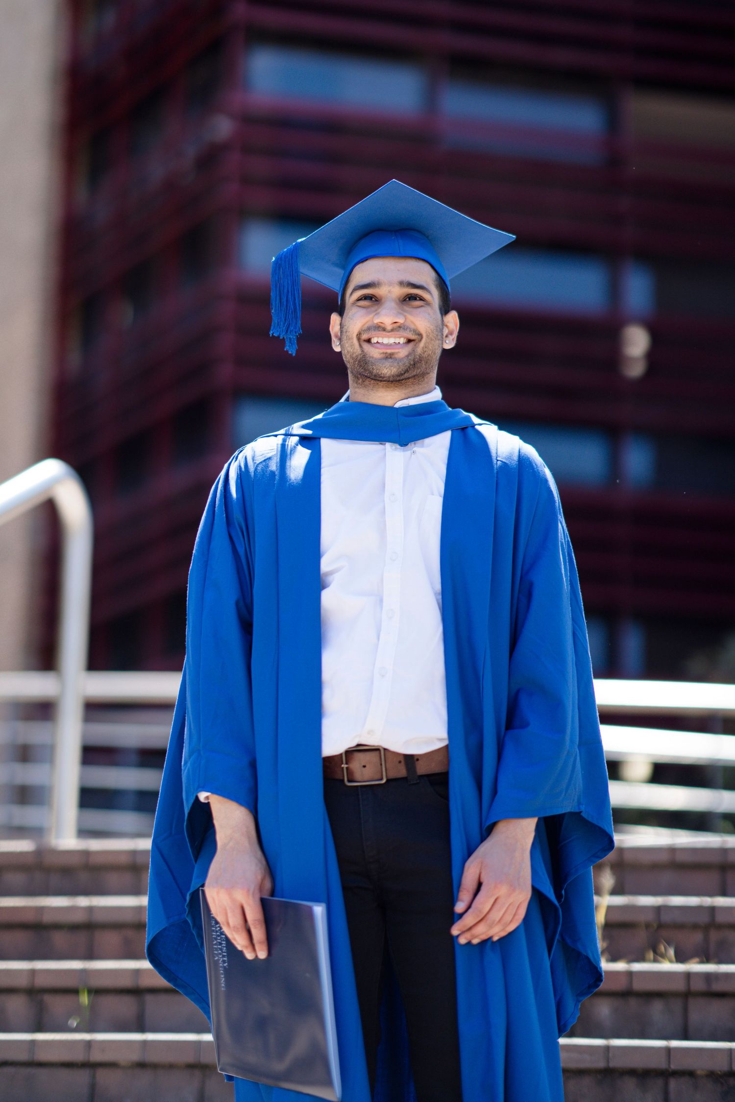 Graduate in blue academic regalia and cap poses on steps with railing.