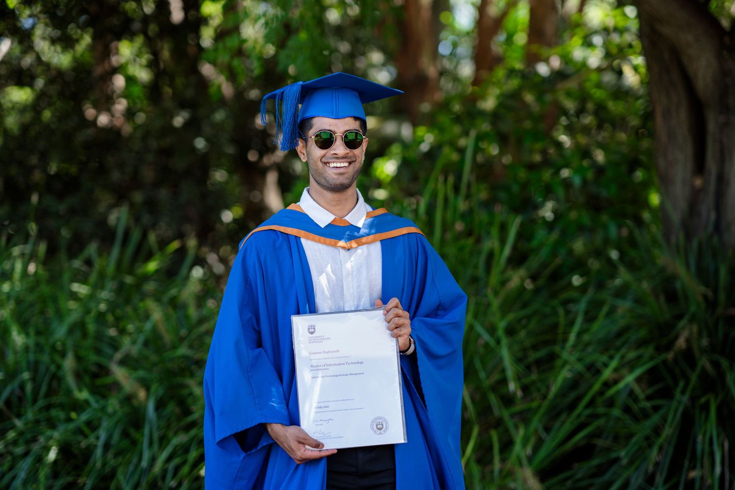 Graduate in academic regalia holds diploma while posing in garden setting.