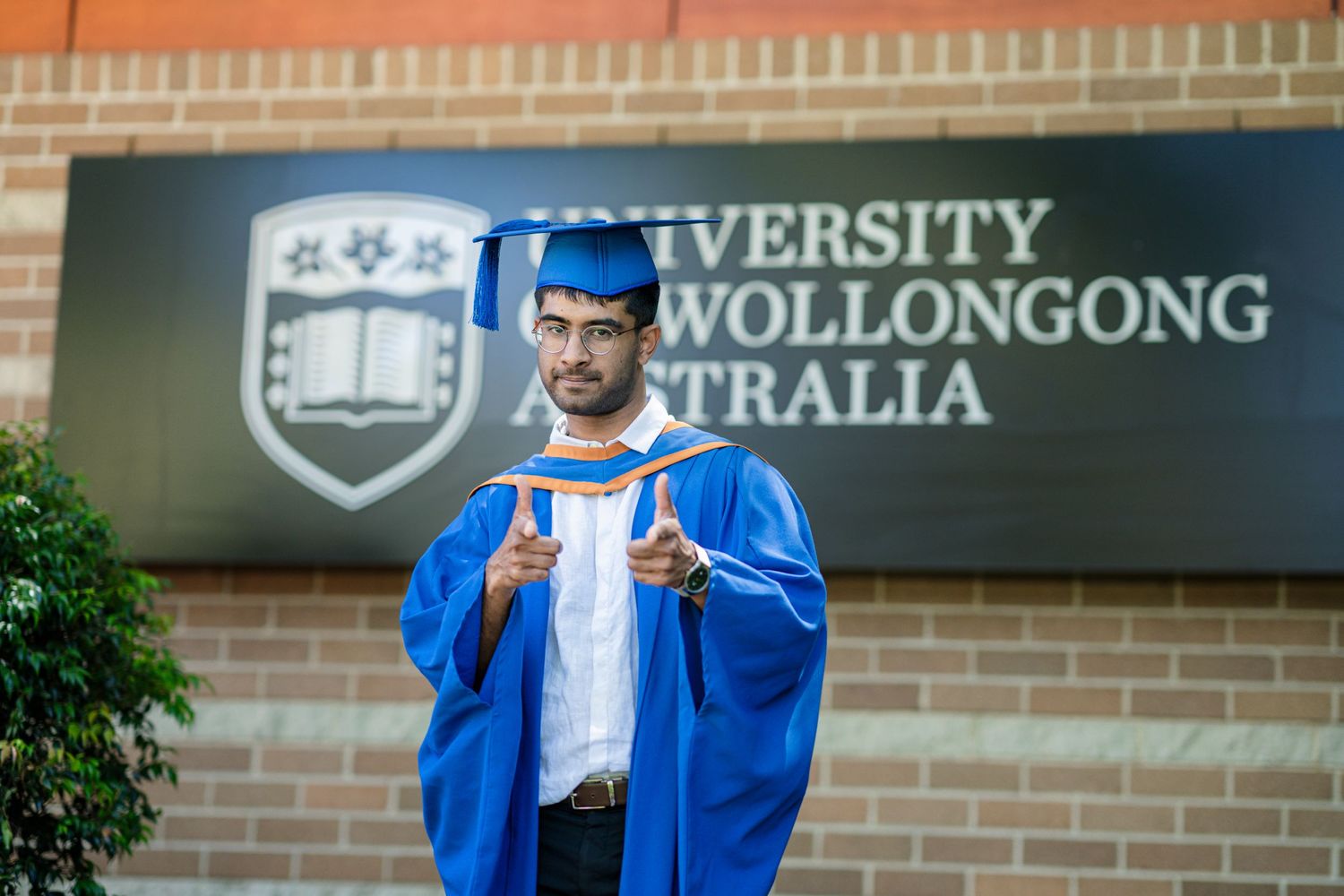 Graduate in blue academic regalia holding diploma in front of University of Wollongong sign.