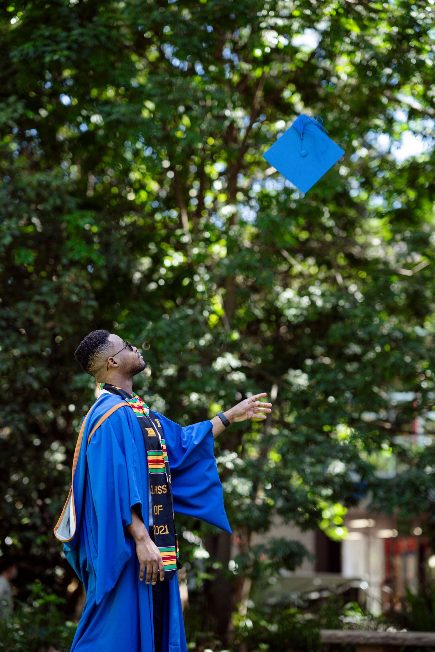 Graduate celebrates outdoors by tossing blue graduation cap with green trees in background.