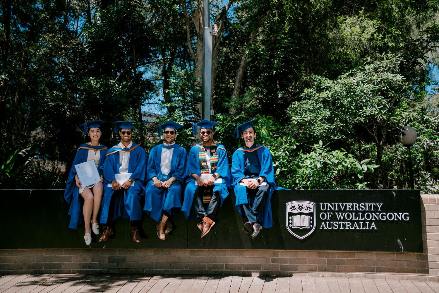 Row of graduates in blue gowns pose on wooden deck with university branding visible.
