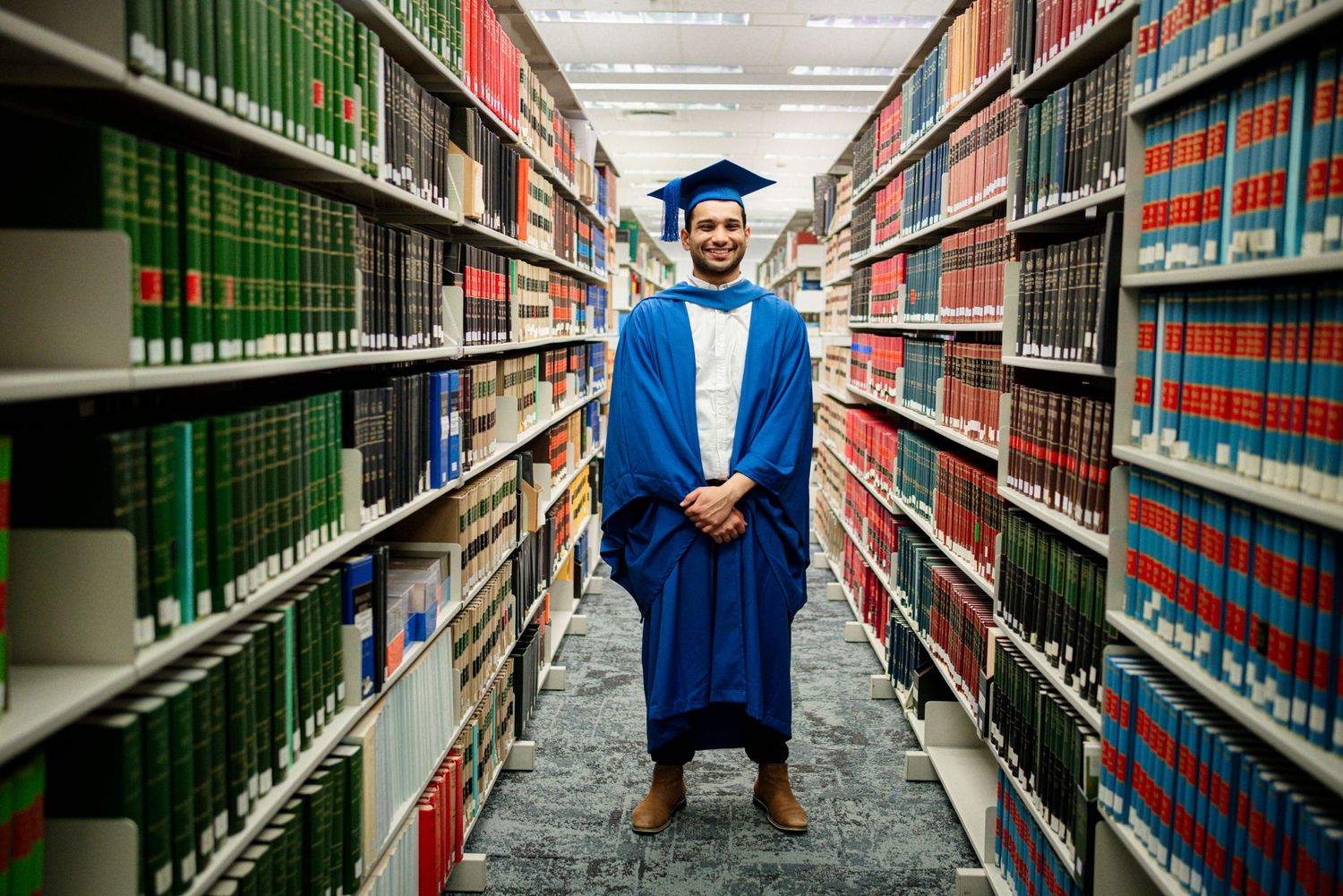 Graduate in blue commencement attire stands confidently in university library stacks.