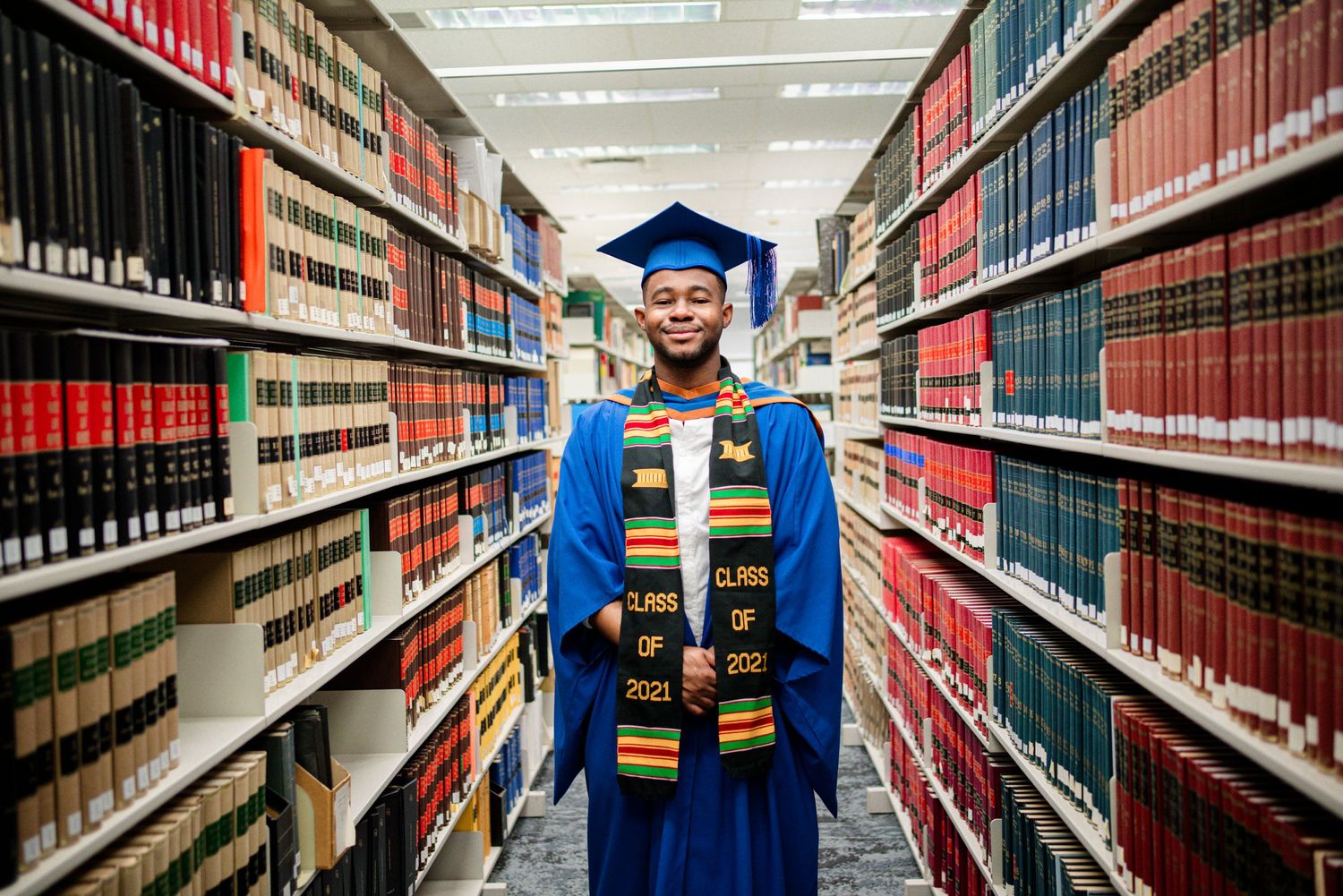 Graduate in blue academic dress poses between tall library shelves filled with books.