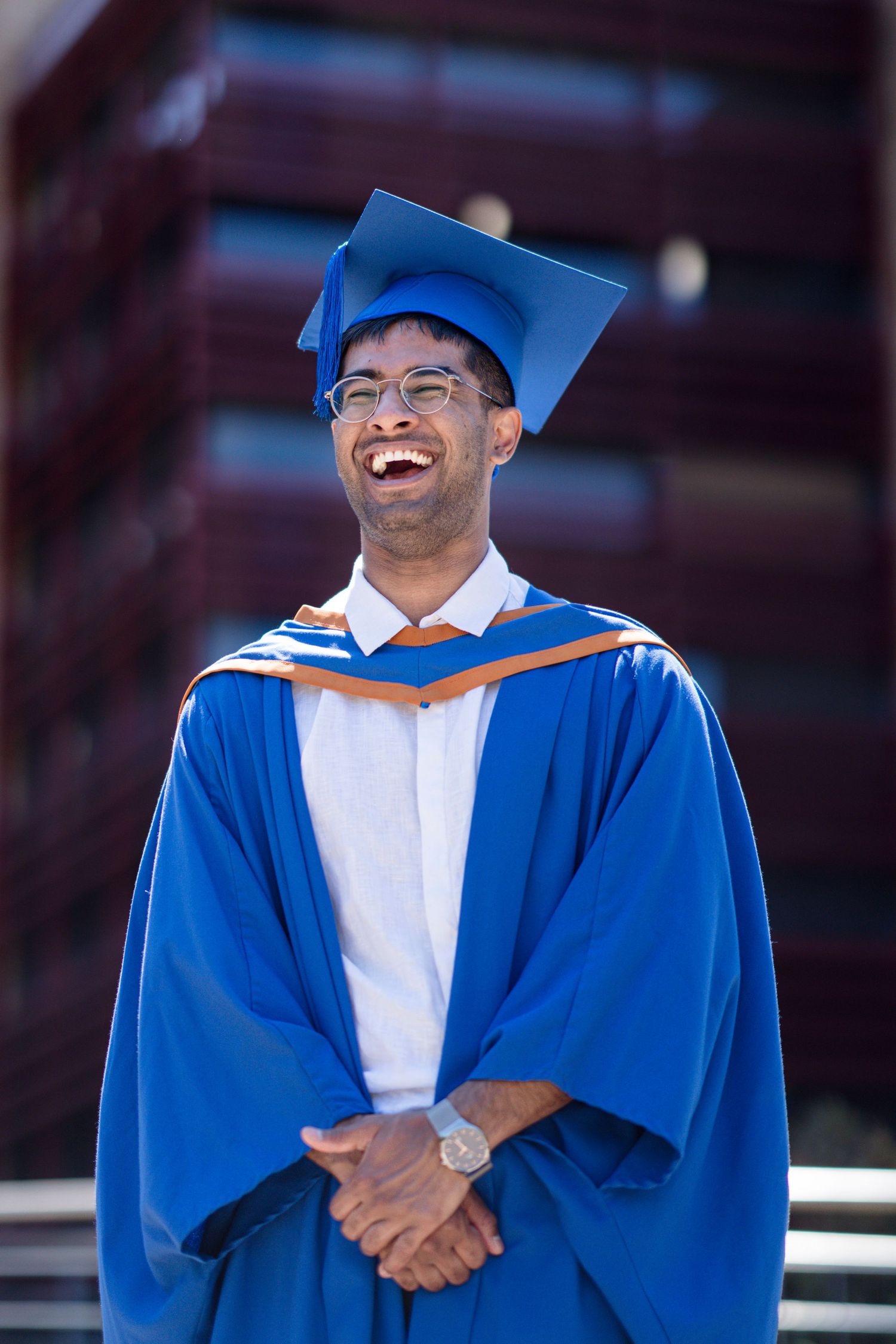 Graduate in blue academic regalia smiles brightly during outdoor photo session.