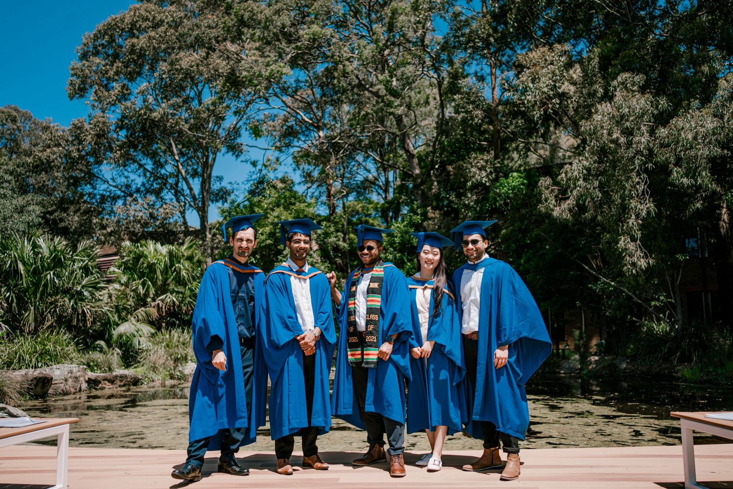 Graduates stand together wearing bright blue graduation gowns against a leafy outdoor backdrop.