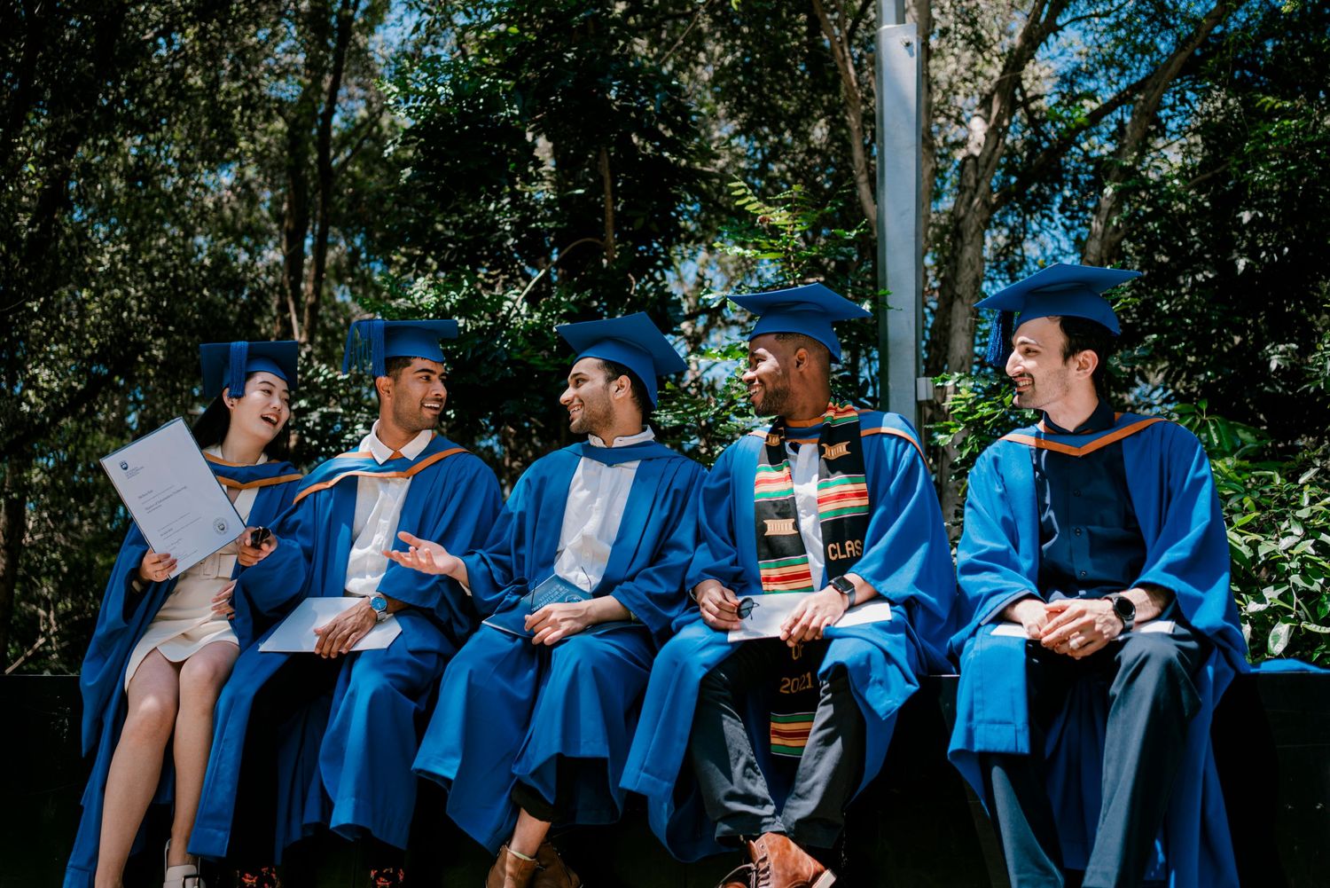 Graduates in blue academic dress share laughs while seated on wooden platform.