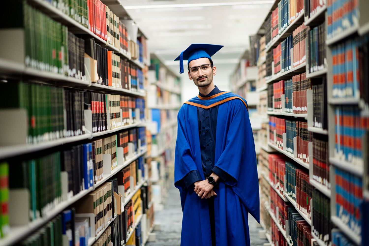 Graduate wearing blue commencement gown photographed in library with rows of books in background.