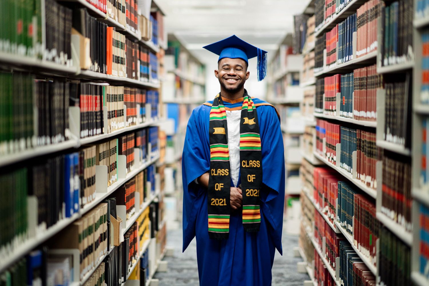 Graduate in blue academic regalia and stoles poses in university library between stacks of books.