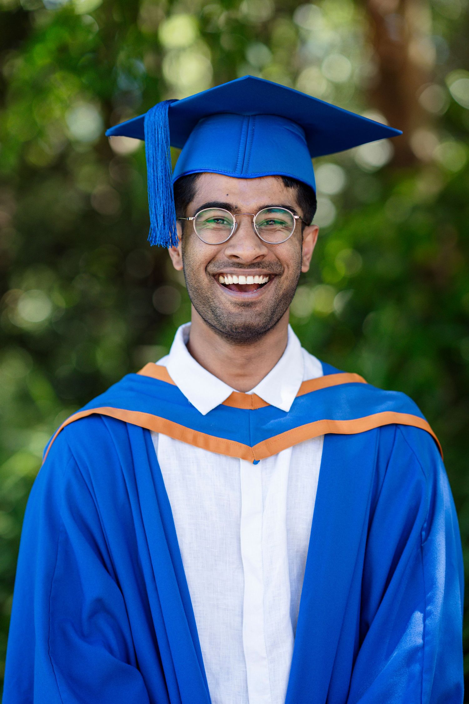 Graduate in blue academic regalia and white stole photographed outdoors with blurred green foliage background.