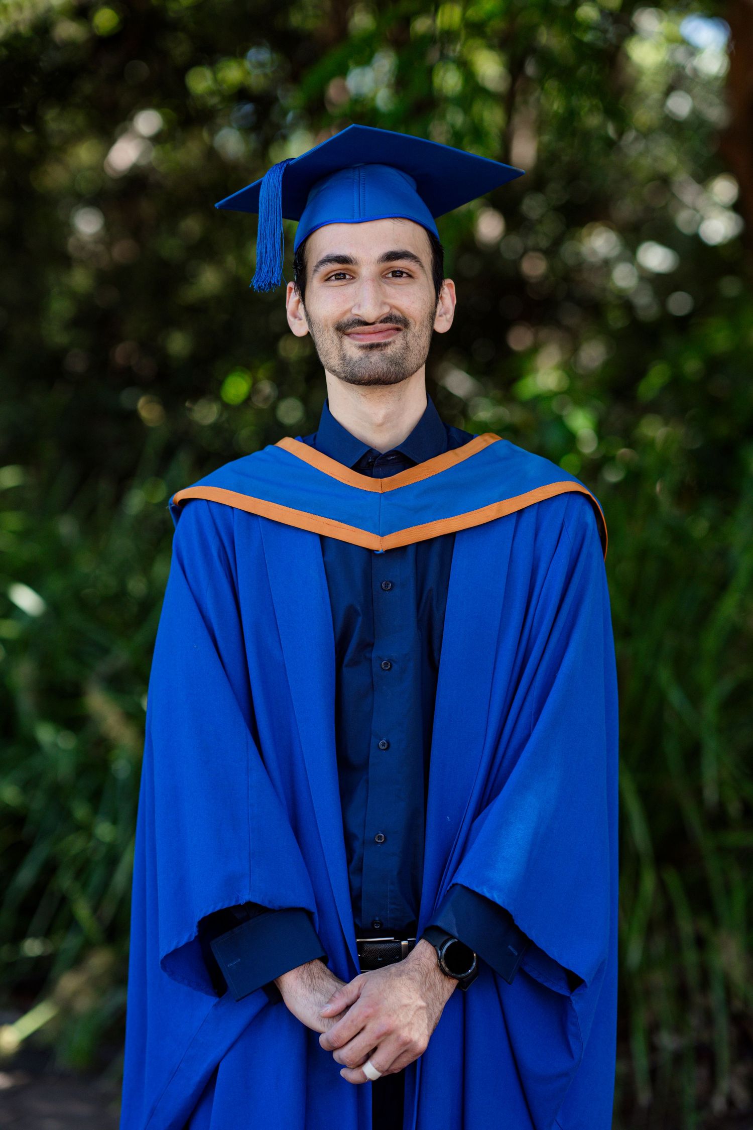 Graduate wearing traditional blue commencement regalia poses in front of green landscaping.