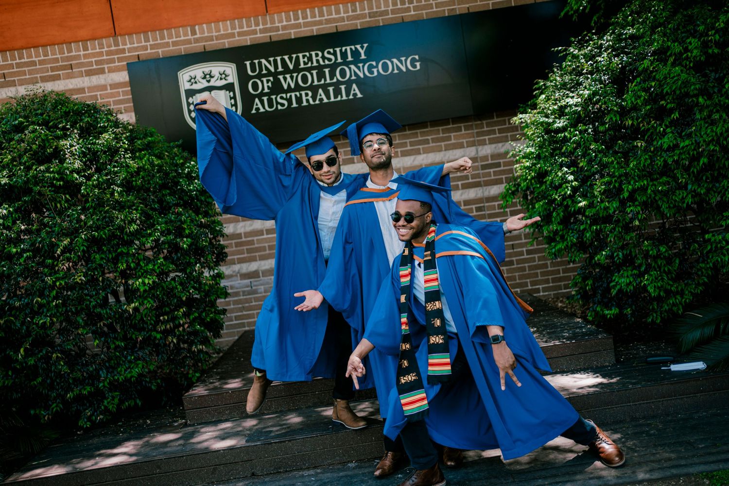 Graduates in blue caps and gowns pose playfully on wooden deck at University of Wollongong campus.