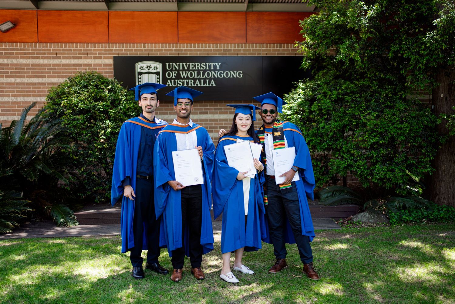 Four graduates in blue academic regalia posing with diplomas outside university building.