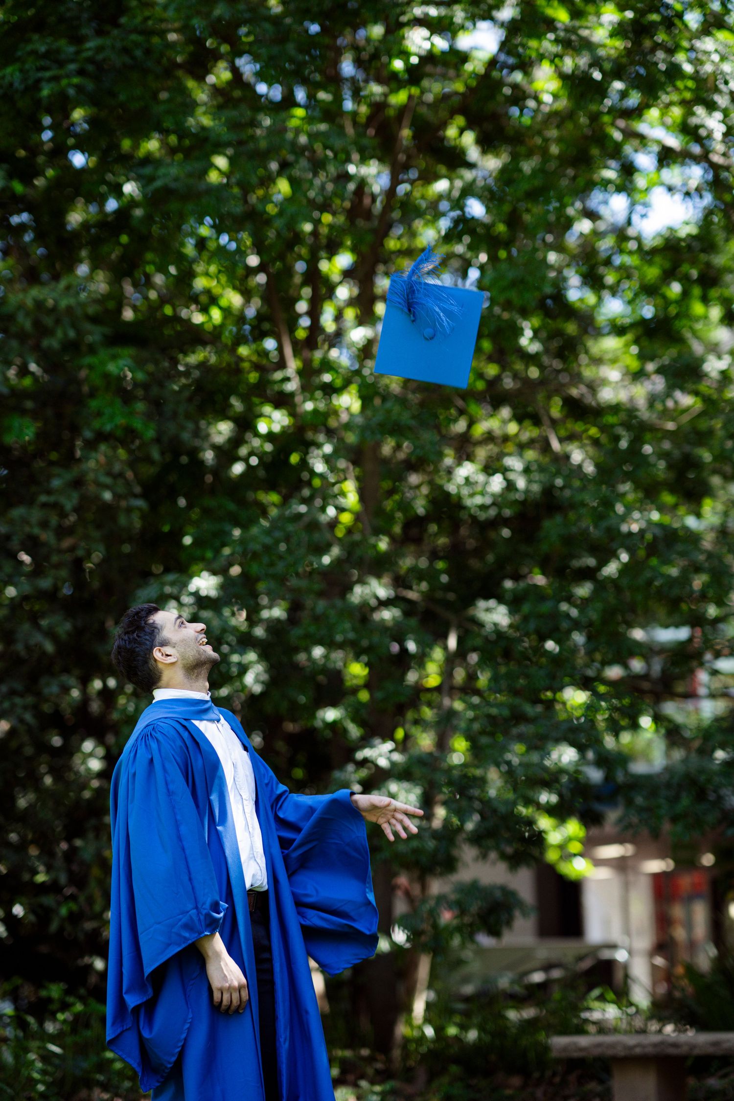 Graduate in traditional blue regalia throws cap in celebratory pose among green trees.