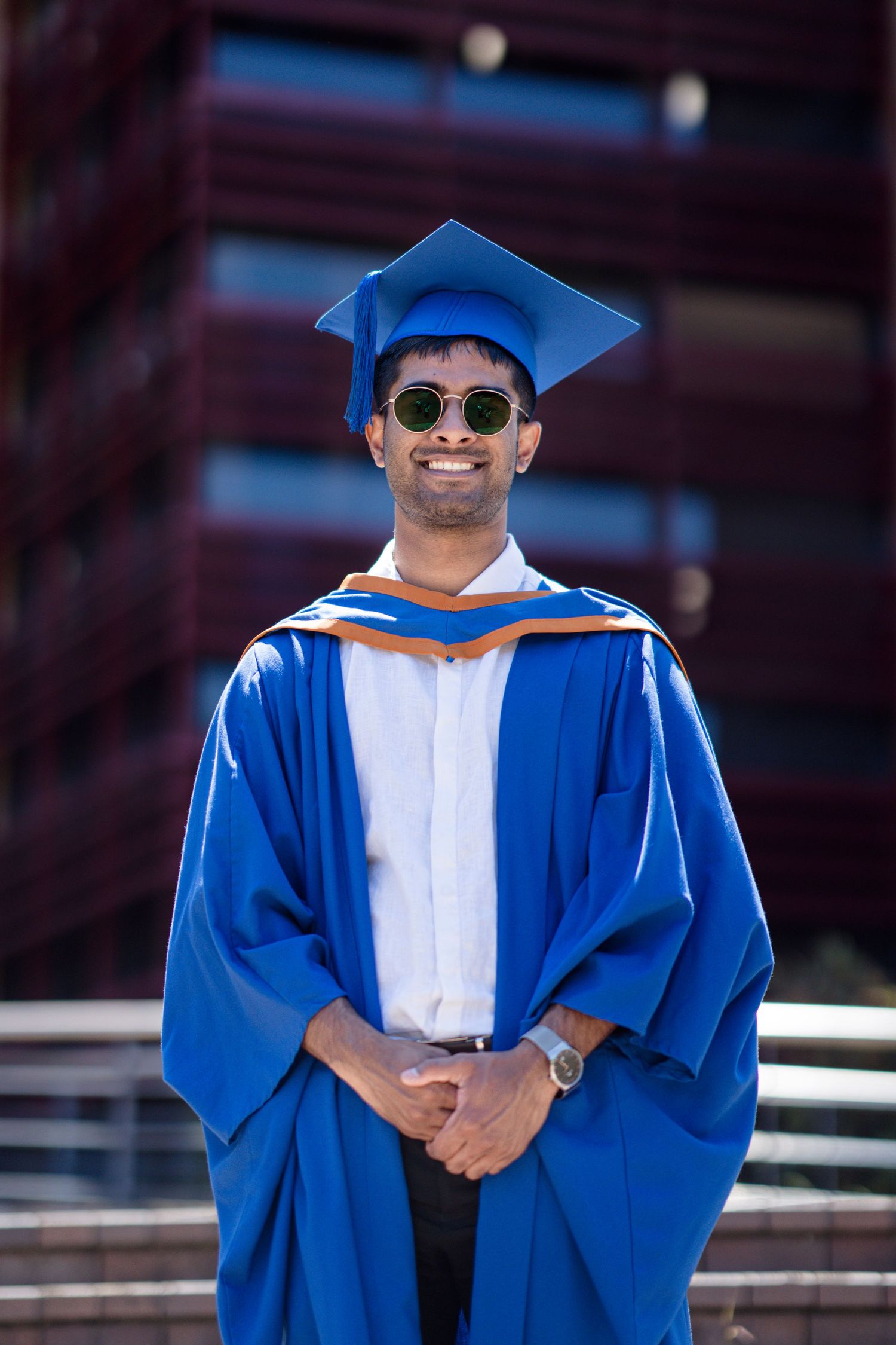 Graduate wearing sunglasses and blue graduation gown poses confidently outdoors.