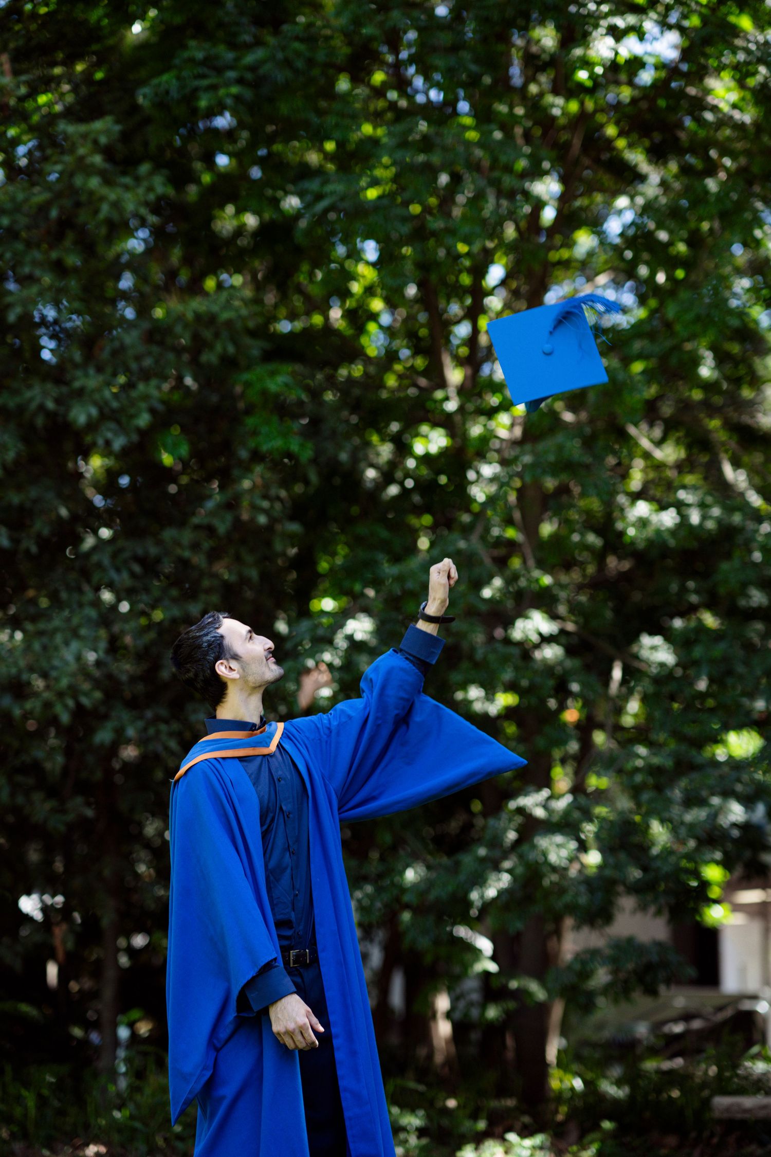 Graduate in blue cap and gown throws graduation cap joyfully in outdoor setting.