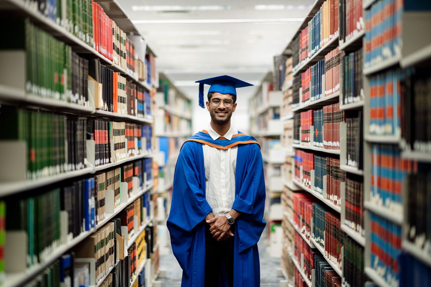 Graduate in blue cap and gown stands between rows of library bookshelves.