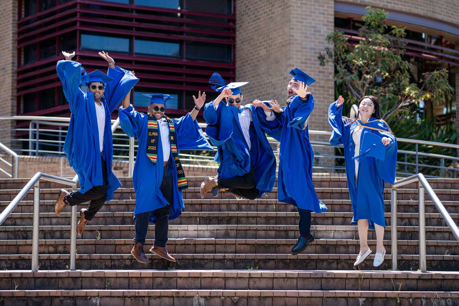 Graduates in blue regalia gather on concrete steps for celebration photos.