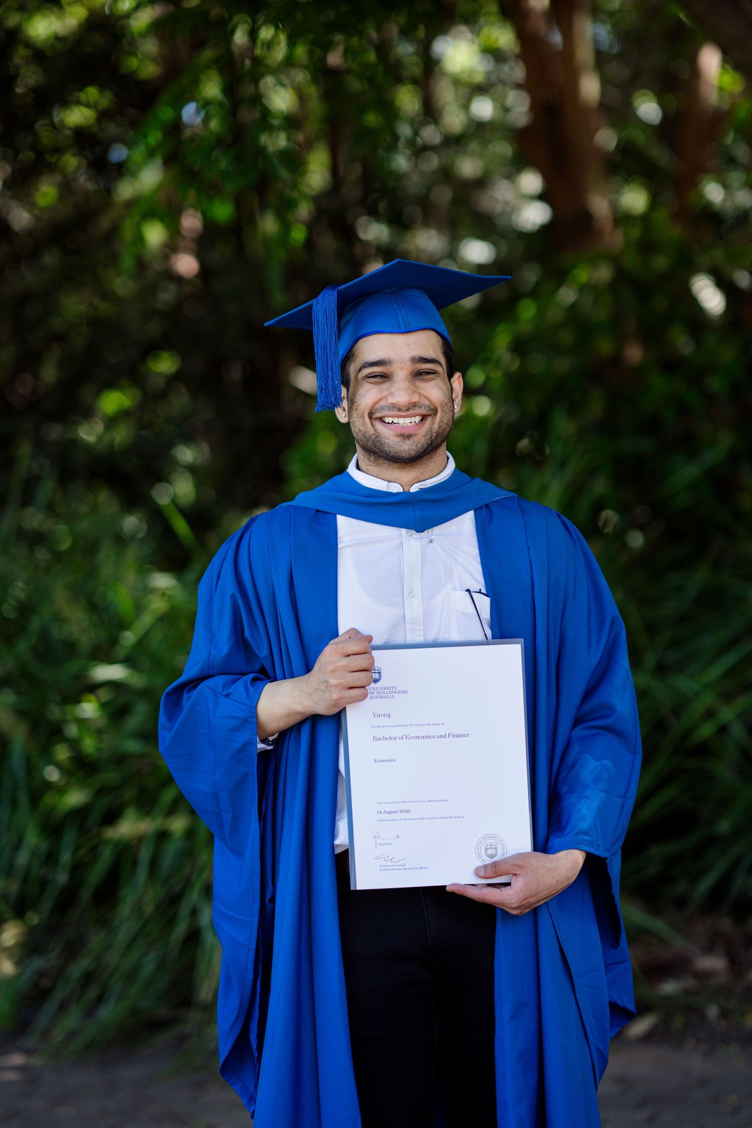 Graduate in blue academic regalia proudly displays diploma while standing in outdoor garden setting.