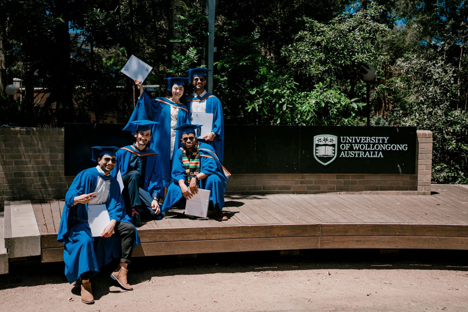 Students in academic regalia celebrate graduation on outdoor wooden platform surrounded by greenery.